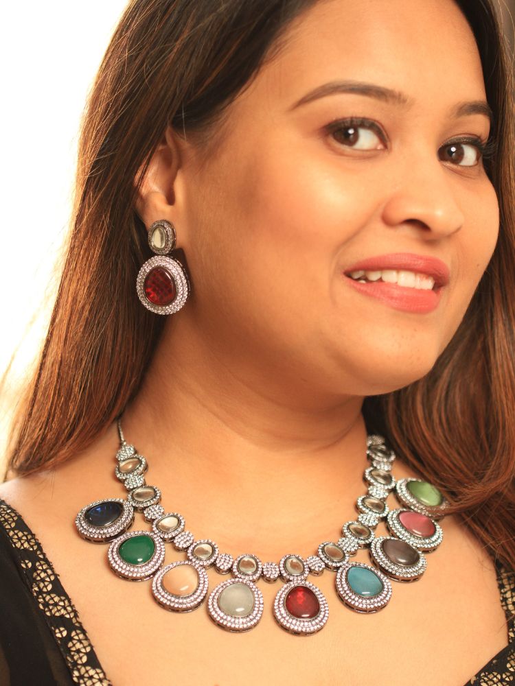 A woman with brown hair down smiles to the side, wearing the Multi-Color Elizabeth Necklace Set featuring a statement Victorian-style necklace and matching earrings with large colorful stones, paired with a sparkly dark top.