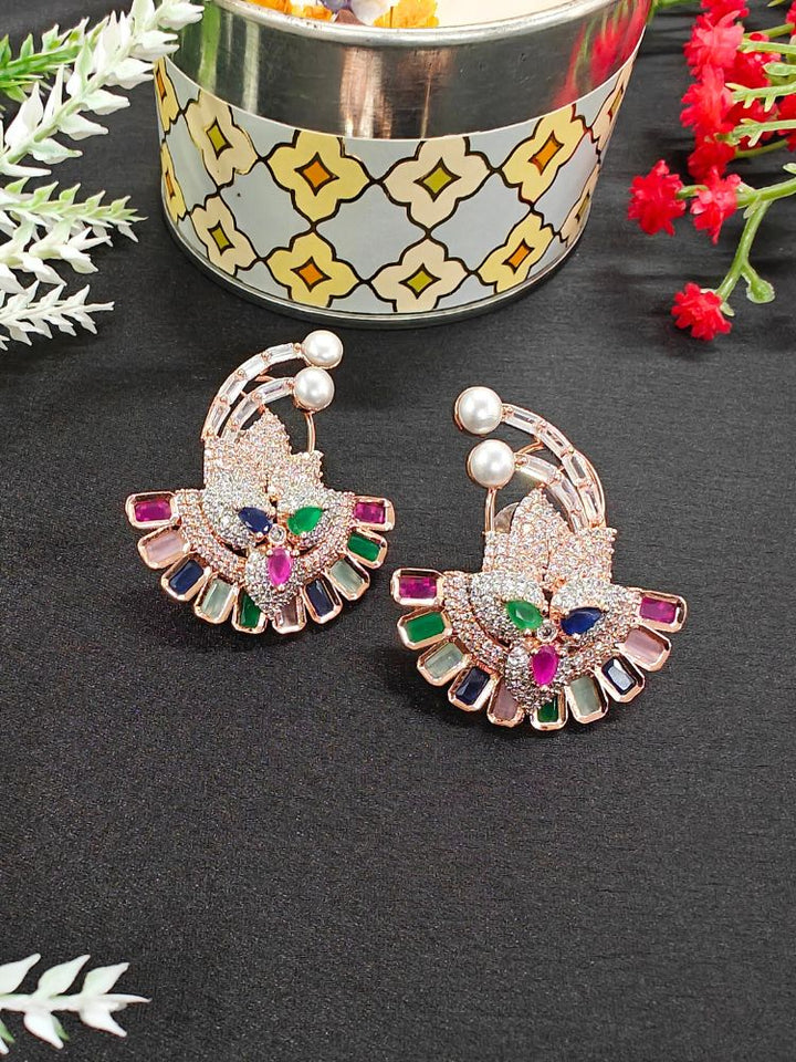 The Multi-color Hansini Earrings feature an ornate floral design with American diamonds and pearls, displayed on a black surface near a decorative container and red flowers—perfect for pairing with your favorite cocktail gown.