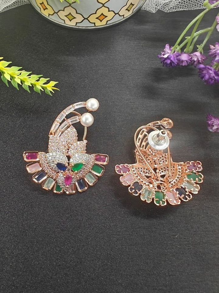 The Multi-color Hansini Earrings feature an ornate floral design with American diamonds and pearls, displayed on a black surface near a decorative container and red flowers—perfect for pairing with your favorite cocktail gown.