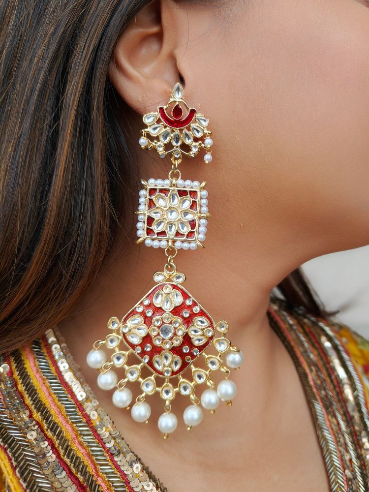 A close-up of a womans ear wearing Myra Earrings—large, ornate Meenakari earrings with red, gold design, surrounded by white pearls, studded with Kundan, pearls and gemstones—complementing her traditional yellow and gold attire.