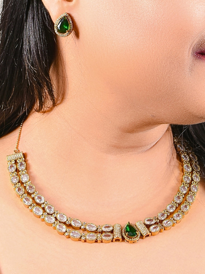 A woman smiles with glossy lips, wearing the Nairika Green Necklace Set—a gold necklace featuring American Diamonds and a vivid Green center stone—paired with matching colored stone stud earrings.