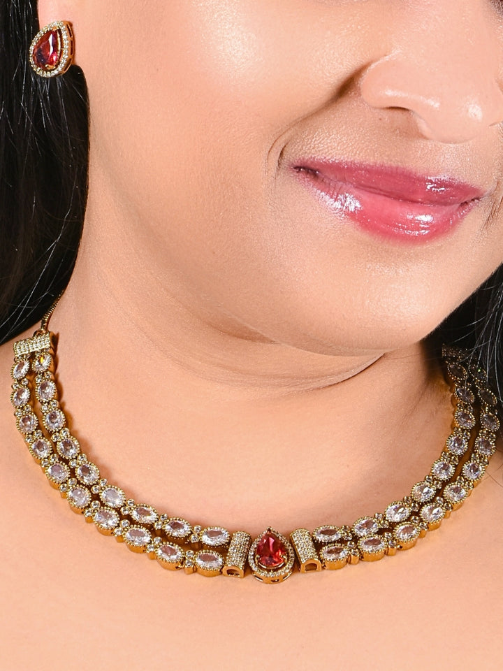 A woman smiles with glossy lips, wearing the Nairika Red Necklace Set—a gold necklace featuring American Diamonds and a vivid red center stone—paired with matching colored stone stud earrings.