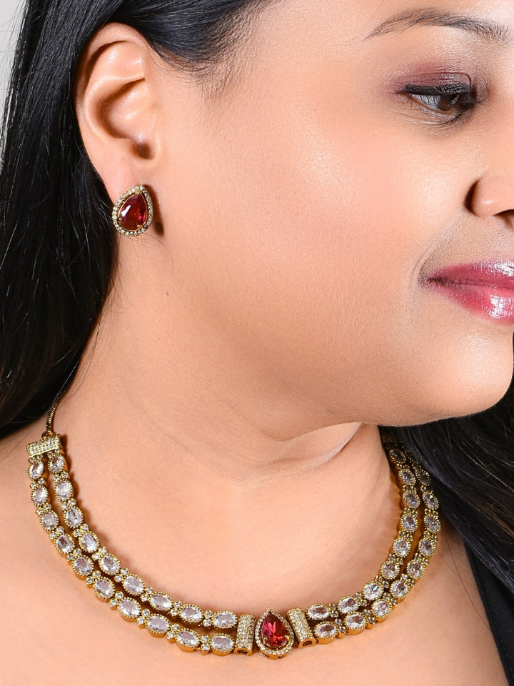 A woman smiles with glossy lips, wearing the Nairika Red Necklace Set—a gold necklace featuring American Diamonds and a vivid red center stone—paired with matching colored stone stud earrings.