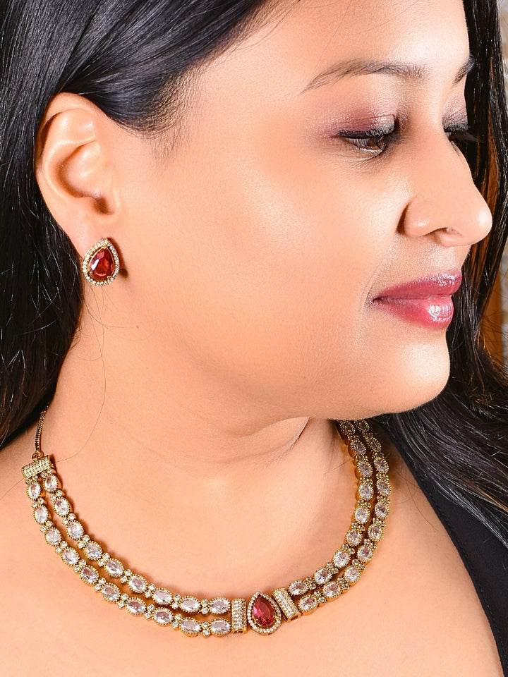 A woman smiles with glossy lips, wearing the Nairika Red Necklace Set—a gold necklace featuring American Diamonds and a vivid red center stone—paired with matching colored stone stud earrings.