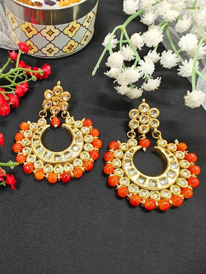 Natalia Orange Earrings—crescent moon-shaped chandbali, gold-toned Kundan stones and orange beads—ideal for Mehendi functions or as wedding ceremony accessories shown on a black background with red and white flowers. 