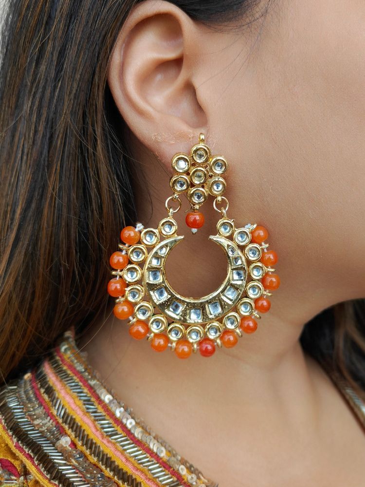 A close-up of a womans ear wearing Natalia Orange Earrings—crescent moon-shaped chandbali, gold-toned Kundan stones and purple beads—ideal for Mehendi functions or as wedding ceremony accessories.