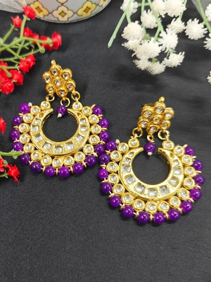Natalia Purple Earrings—crescent moon-shaped chandbali, gold-toned Kundan stones and purple beads—ideal for Mehendi functions or as wedding ceremony accessories, shown on a black ground with red and white flowers. 