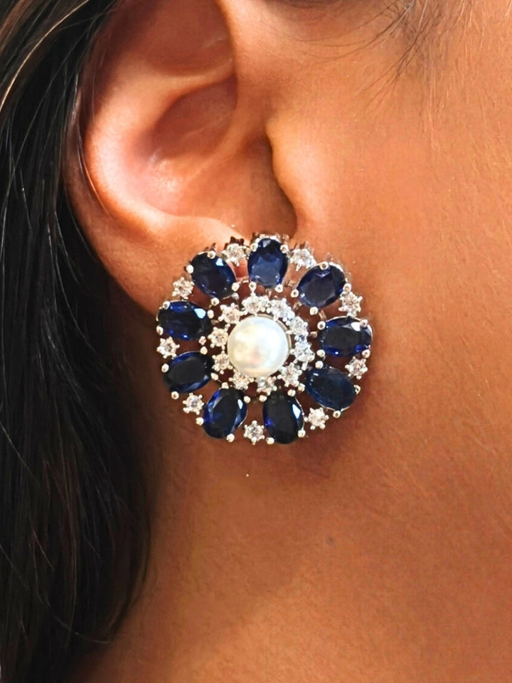 A close-up of a womans ear wearing the Navy Blue Blossom stud Earrings—large, circular Navy Blue earrings with a pearl at the center, colorful accents, and a border of sparkling American Diamonds. The stud earrings give a flower earrings feel. 
