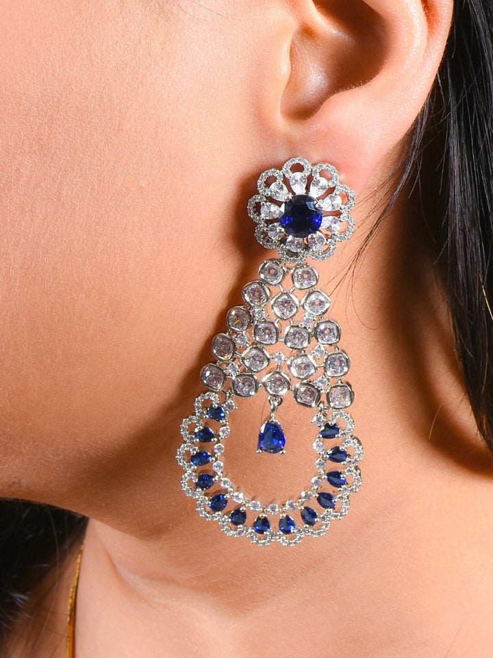 A close-up of a womans ear features the Navy Blue Darcie Earrings, designed with American Diamonds in floral and circular patterns and finished with a dangling blue teardrop gem—ideal for complementing a cocktail gown.
