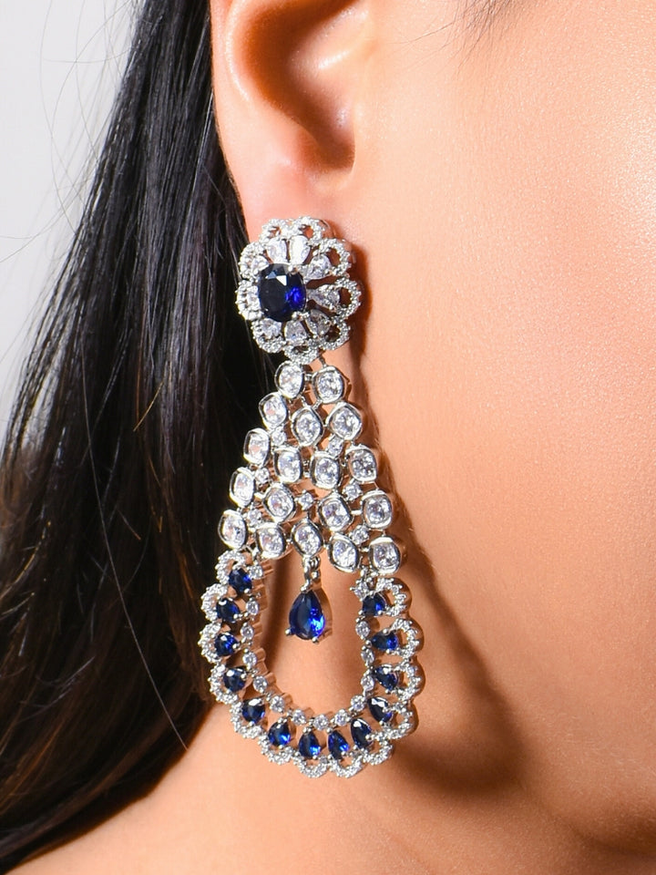 A close-up of a womans ear features the Navy Blue Darcie Earrings, designed with American Diamonds in floral and circular patterns and finished with a dangling blue teardrop gem—ideal for complementing a cocktail gown.
