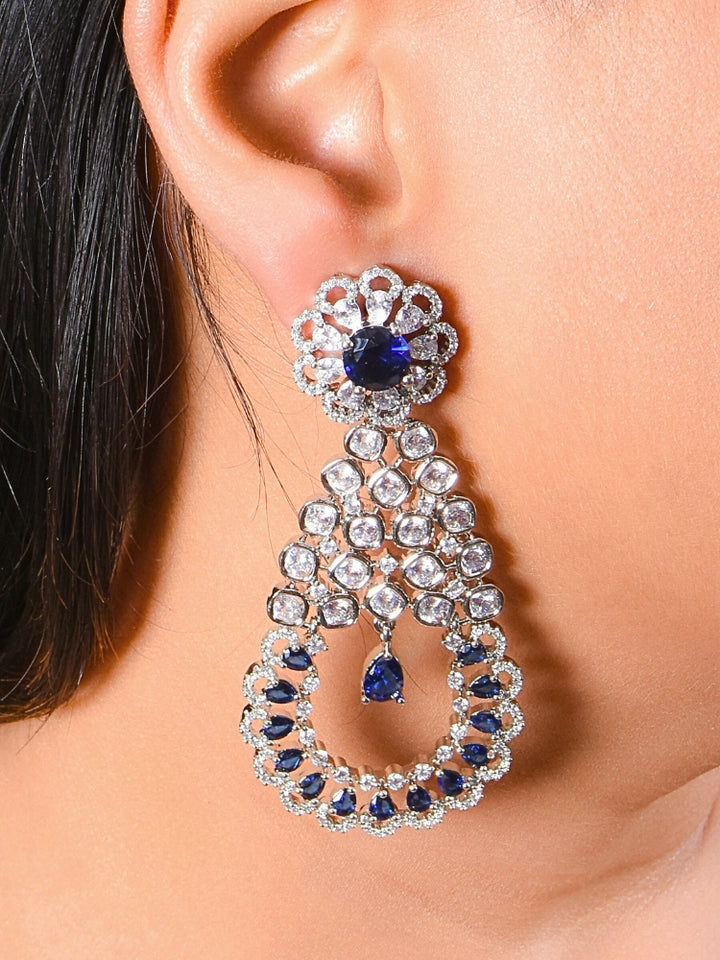 A close-up of a womans ear features the Navy Blue Darcie Earrings, designed with American Diamonds in floral and circular patterns and finished with a dangling blue teardrop gem—ideal for complementing a cocktail gown.