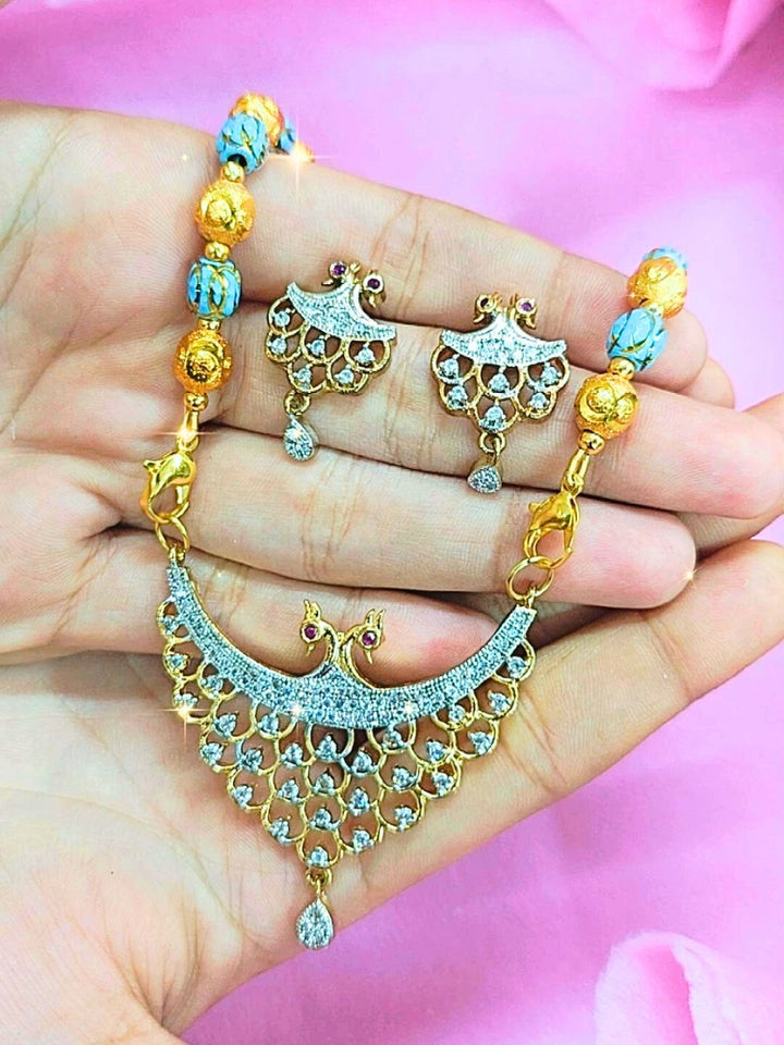 A hand holds the Niharika Mangalsutra—a gold necklace with black mangalsutra beads and matching earrings, pendant featuring sparkling American Diamonds—set against a soft pink background, perfect for your wedding ceremony dress. Diamond mangalsutra. Modern mangalsutra. 