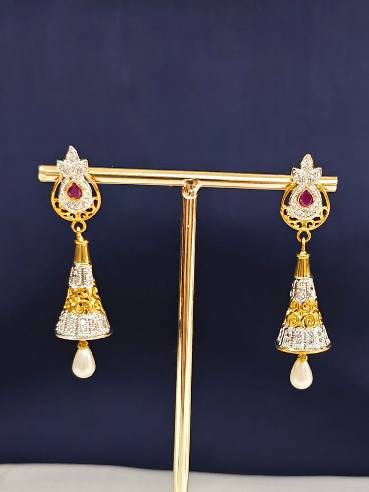 The Niral Earrings are ornate gold and silver jhumkas featuring red gemstones, intricate detailing, and pearl drops. Crafted in brass, they are displayed on a gold stand against a dark blue background.