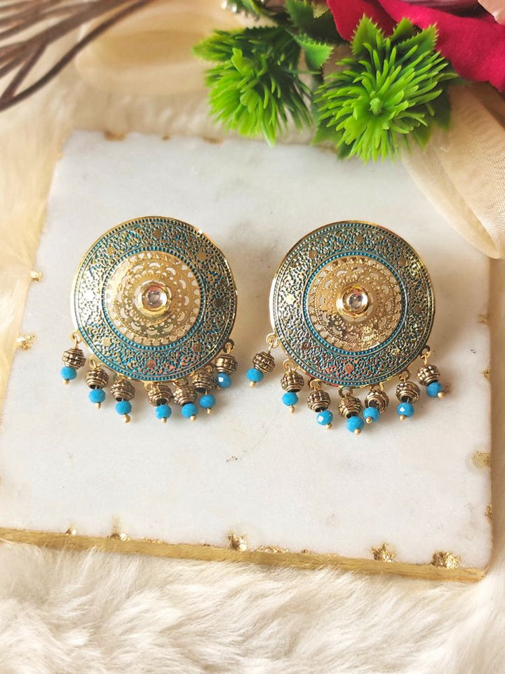 The Nishtha Earrings are round blue earrings with ornate gold and teal patterns in meenakari and filigree design, along with small blue beads, ideal as an ethnic accessory for a wedding outfit. Displayed on white marble with green artificial plants in the background.