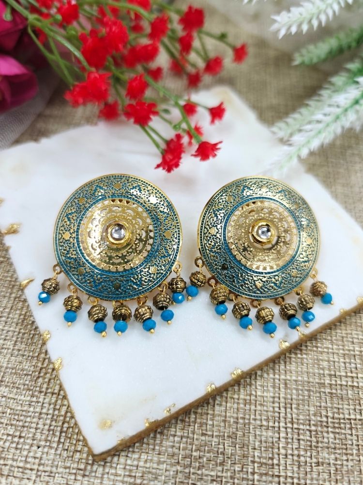 The Nishtha Earrings are round blue earrings with ornate gold and teal patterns in meenakari and filigree design, along with small blue beads, ideal as an ethnic accessory for a wedding outfit. Displayed on white marble with Red artificial Flower in the background.