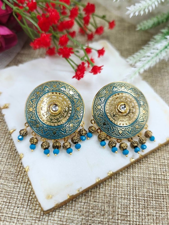 The Nishtha Earrings are round blue earrings with ornate gold and teal patterns in meenakari and filigree design, along with small blue beads, ideal as an ethnic accessory for a wedding outfit. Displayed on white marble with Red artificial Flower in the background.