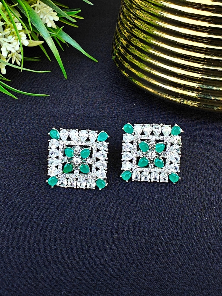 Nyra Green Earrings: Square-shaped earrings featuring clear American Diamonds and green emerald stones in an intricate pattern, displayed on dark fabric near green leaves and a gold accent.