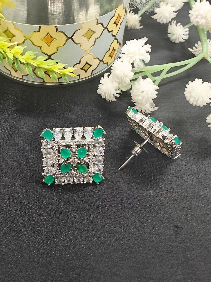 Nyra Green Earrings: Square-shaped earrings featuring clear American Diamonds and green emerald stones in an intricate pattern, displayed on dark fabric near green leaves and a gold accent.