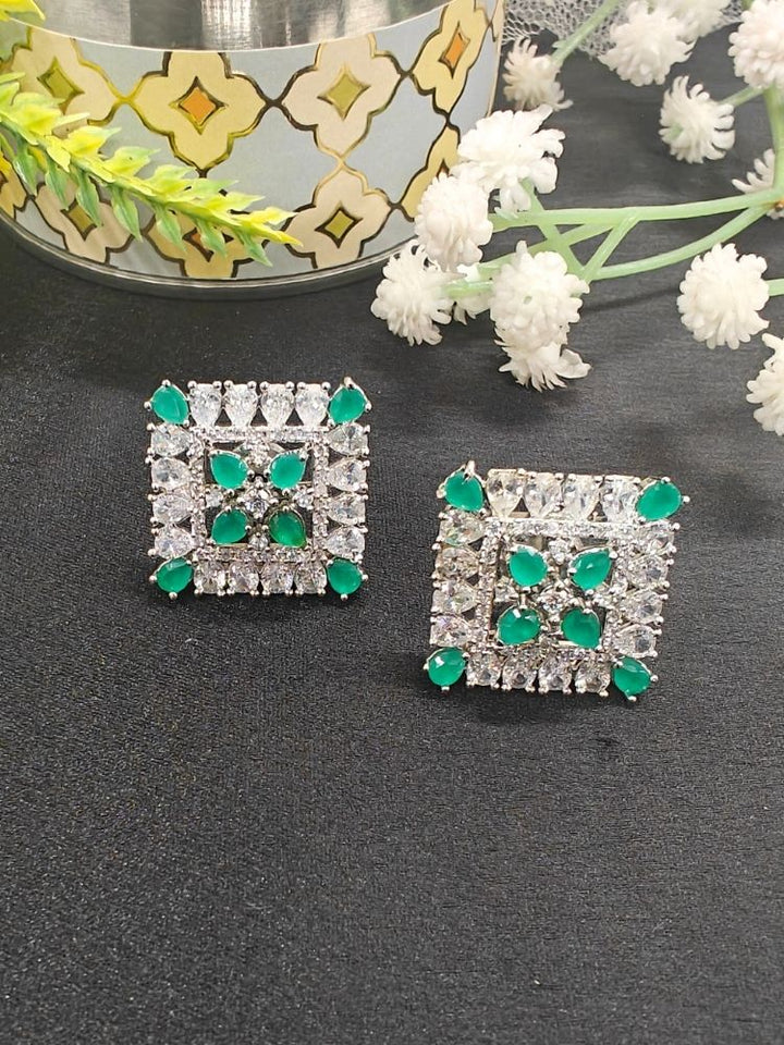 Nyra Green Earrings: Square-shaped earrings featuring clear American Diamonds and green emerald stones in an intricate pattern, displayed on dark fabric near green leaves and a gold accent.
