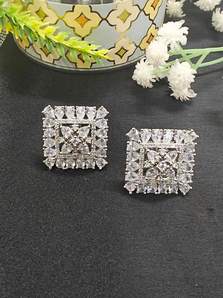 Nyra White Earrings: Square-shaped earrings featuring clear American Diamonds and White stones in an intricate pattern, displayed on dark fabric near green leaves and a gold accent.