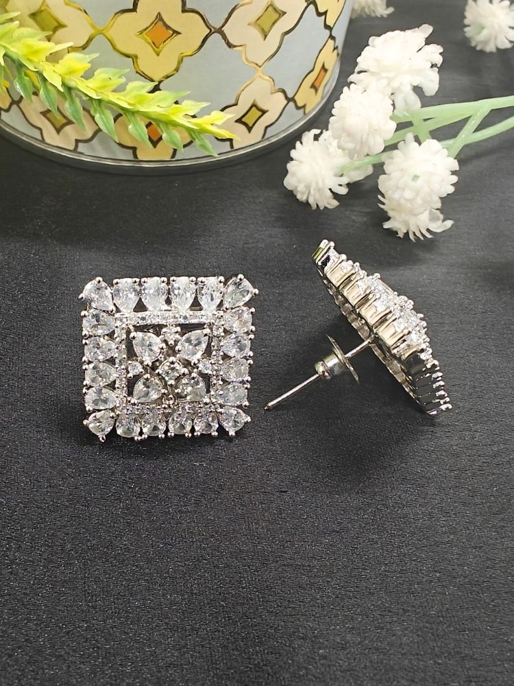 Nyra White Earrings: Square-shaped earrings featuring clear American Diamonds and White stones in an intricate pattern, displayed on dark fabric near green leaves and a gold accent.