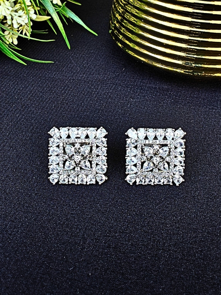Nyra White Earrings: Square-shaped earrings featuring clear American Diamonds and White stones in an intricate pattern, displayed on dark fabric near green leaves and a gold accent.