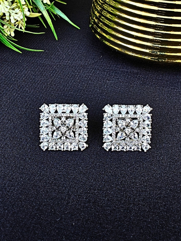 Nyra White Earrings: Square-shaped earrings featuring clear American Diamonds and White stones in an intricate pattern, displayed on dark fabric near green leaves and a gold accent.