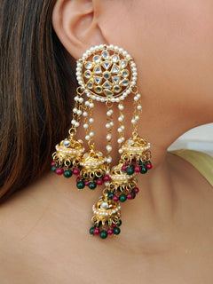 A close-up of a womans ear wearing Ojasvi Red & Green Earrings, featuring intricate gold Kundan work surrounded by pearls, also with pearls holding on to five small kundan jhumkis with red and green beads that cover her ear and extend below it.