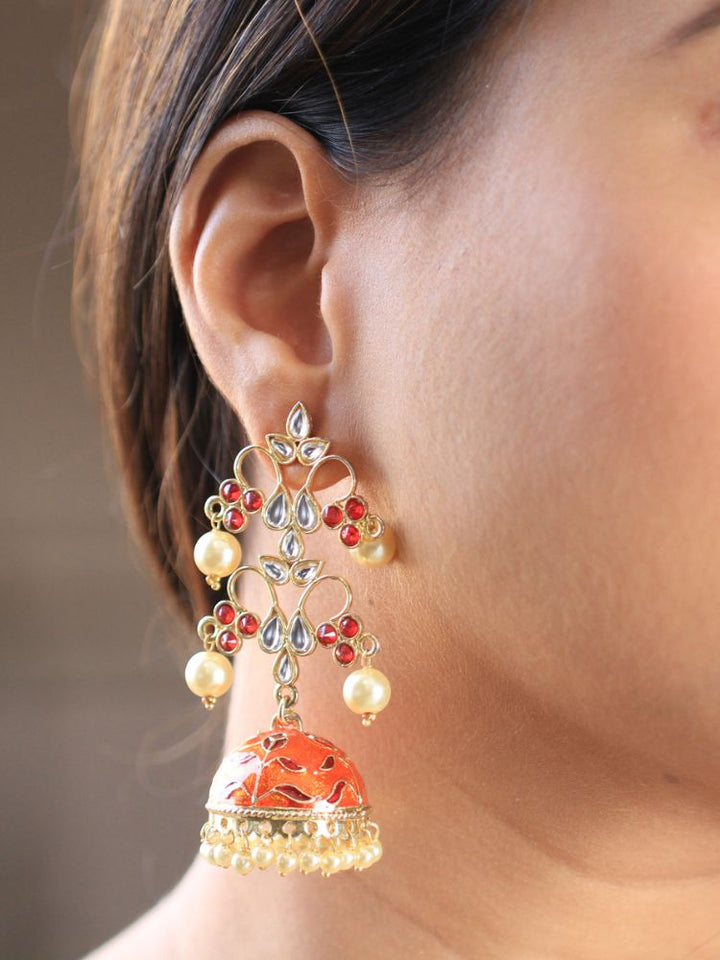 A close-up of a woman’s ear showcases the Orange Aisha Long Jhumka—an ornate, gold-tone earring with red colored stones, along with kundan and and pearl work on top, along with an orange jhumka with Meenakari work adorned with dangling pearls.
