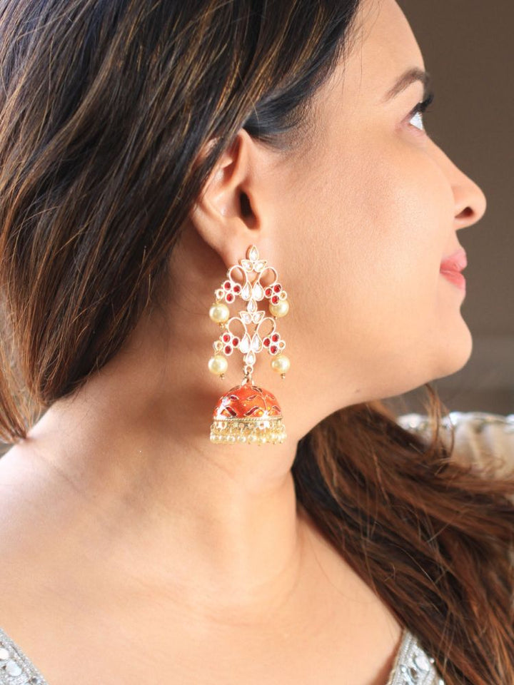 A close-up of a woman’s ear showcases the Orange Aisha Long Jhumka—an ornate, gold-tone earring with red colored stones, along with kundan and and pearl work on top, along with an orange jhumka with Meenakari work adorned with dangling pearls.