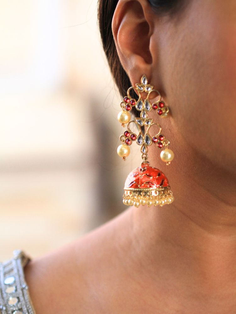 A close-up of a woman’s ear showcases the Orange Aisha Long Jhumka—an ornate, gold-tone earring with red colored stones, along with kundan and and pearl work on top, along with an orange jhumka with Meenakari work adorned with dangling pearls.
