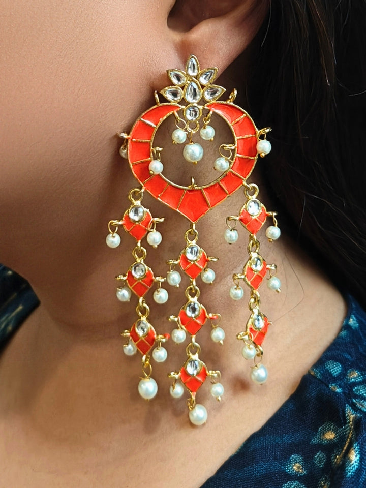A close-up of a person wearing Orange Ankita Earrings—large, ornate Orange and gold earrings with dangling pearls and gemstones in intricate patterns—standing out against dark hair and a blue patterned top. It has meenakari design all over the earrings, and is perfect for wedding functions. 