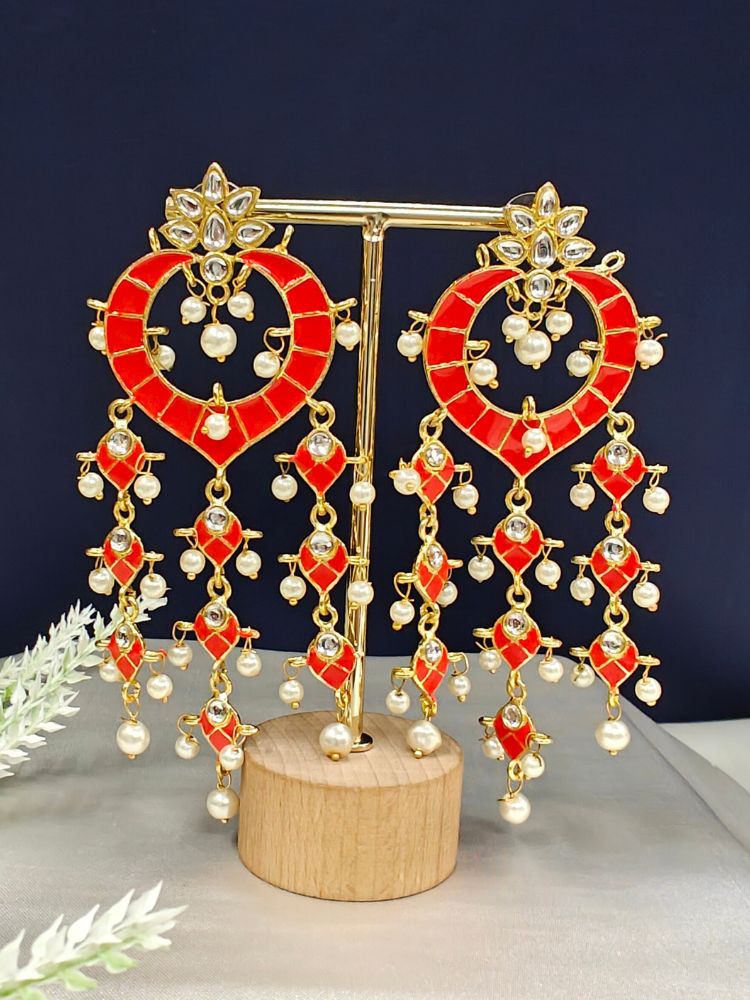 The Orange Ankita Earrings are ornate gold Kundan earrings with Orange enamel, intricate Meenakari work, white stones, and dangling pearl beads, shown on a gold stand with a wooden base against a dark background.
