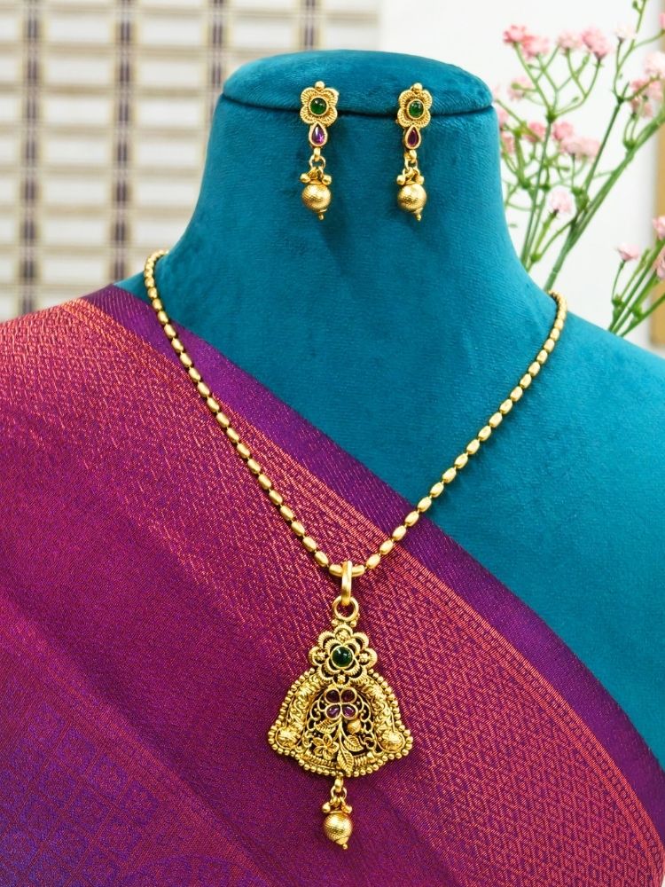 A mannequin displays the Paras Pendant Set— a gold necklace with a large pendant and matching filigree earrings—paired with a purple-maroon saree. Pink flowers decorate the background.