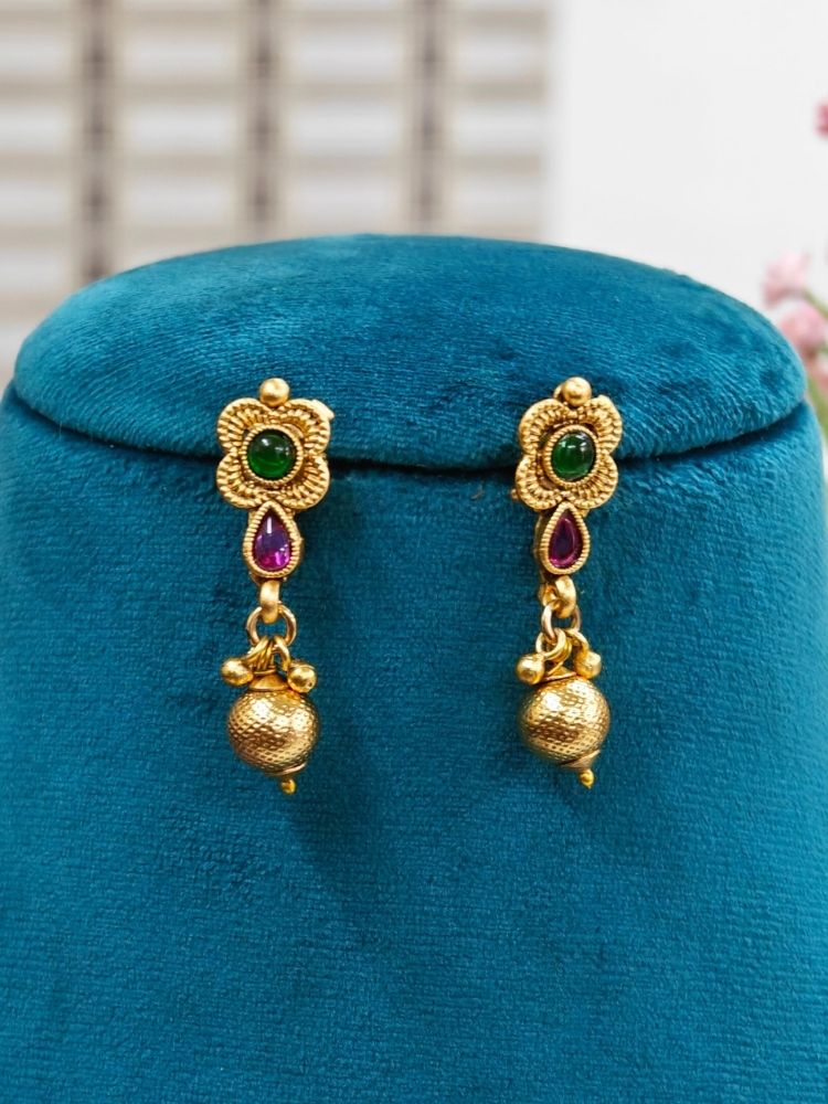 A mannequin displays the Paras Pendant Set— a gold necklace with a large pendant and matching filigree earrings—paired with a purple-maroon saree. Pink flowers decorate the background.