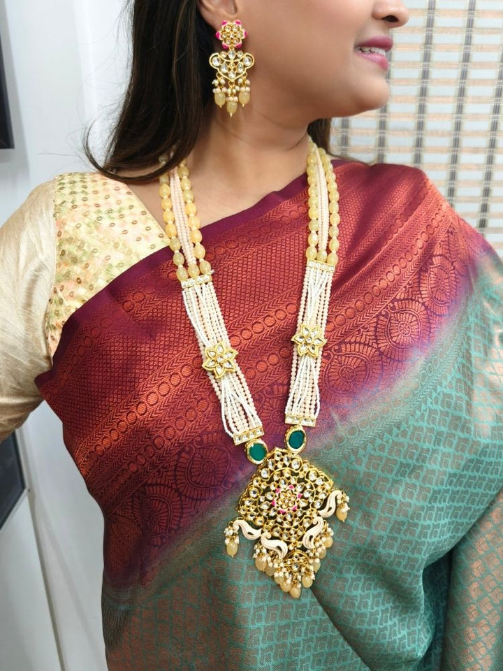 A woman in a cream and maroon-green saree wears the Peach Noor Necklace Set, featuring a long Kundan design with pearls and green stones and matching earrings, as she smiles while looking away from the camera.