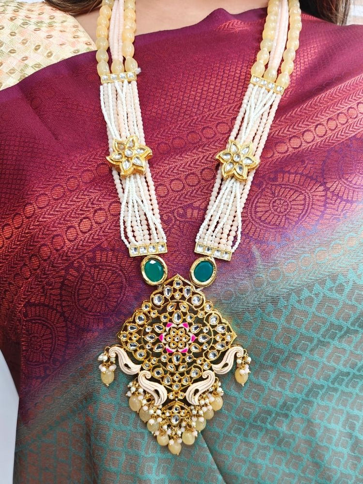 A woman in a cream and maroon-green saree wears the Peach Noor Necklace Set, featuring a long Kundan design with pearls and green stones and matching earrings, as she smiles while looking away from the camera.