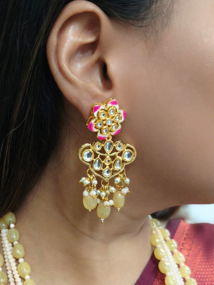 A woman in a cream and maroon-green saree wears the Peach Noor Necklace Set, featuring a long Kundan design with pearls and green stones and matching earrings, as she smiles while looking away from the camera.