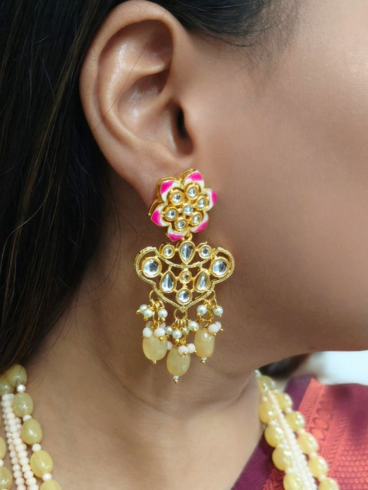A woman in a cream and maroon-green saree wears the Peach Noor Necklace Set, featuring a long Kundan design with pearls and green stones and matching earrings, as she smiles while looking away from the camera.