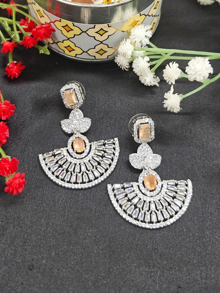 The Peach Nora Earrings feature ornate fan-shaped designs with American Diamonds, displayed on a black surface near red and white flowers and a decorative bowl with floating elements.
