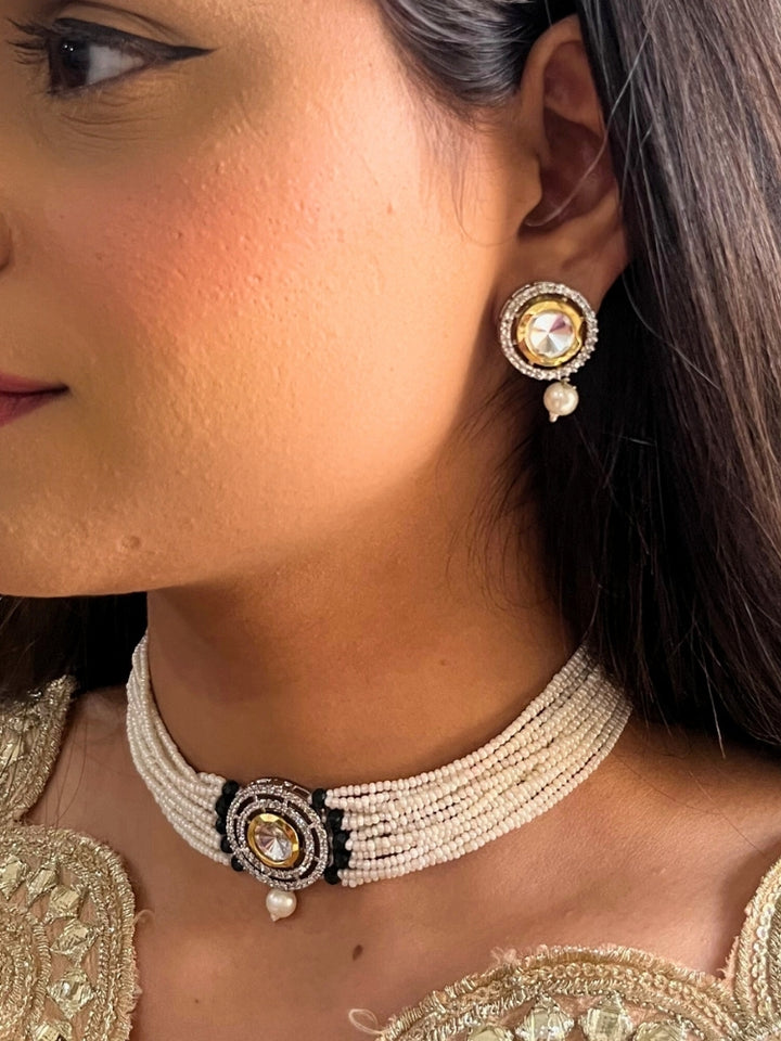 A woman wears the Pearl Black Renu Necklace Set, featuring a multi-strand white pearl choker with Kundan and Black accents, a central jeweled pendant, and matching round earrings adorned with dangling pearls. Kundan choker necklace accentuates her look.