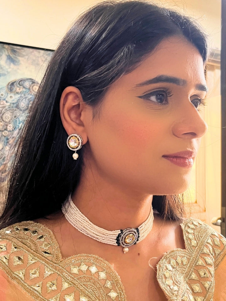 A woman wears the Pearl Black Renu Necklace Set, featuring a multi-strand white pearl choker with Kundan and Black accents, a central jeweled pendant, and matching round earrings adorned with dangling pearls. Kundan choker necklace accentuates her look.