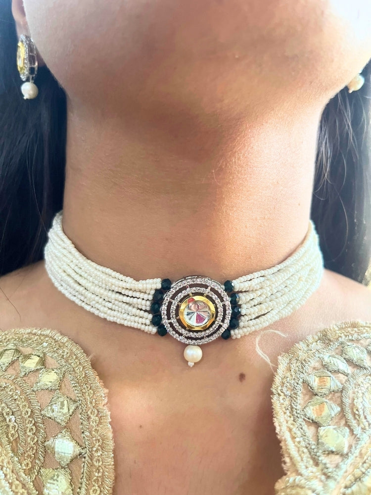 A woman wears the Pearl Black Renu Necklace Set, featuring a multi-strand white pearl choker with Kundan and Black accents, a central jeweled pendant, and matching round earrings adorned with dangling pearls. Kundan choker necklace accentuates her look.