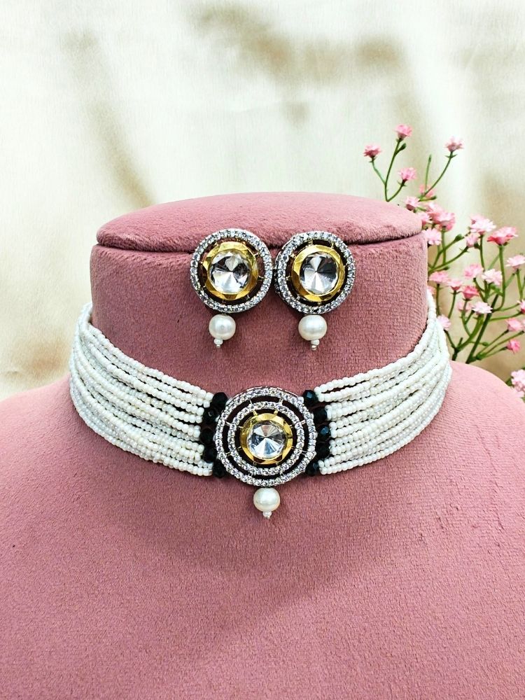 The Pearl Black Renu Necklace Set features a beaded choker with a bold circular pendant and matching stud earrings, embellished with gold, silver, American diamonds, and pearl accents. Displayed on a pink stand with pink flowers in the background.