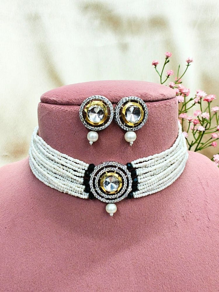 The Pearl Black Renu Necklace Set features a beaded choker with a bold circular pendant and matching stud earrings, embellished with gold, silver, American diamonds, and pearl accents. Displayed on a pink stand with pink flowers in the background.