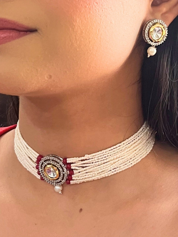 A woman wears the Pearl Red Renu Necklace Set, featuring a multi-strand white pearl choker with Kundan and red accents, a central jeweled pendant, and matching round earrings adorned with dangling pearls. Kundan choker necklace accentuates her look.