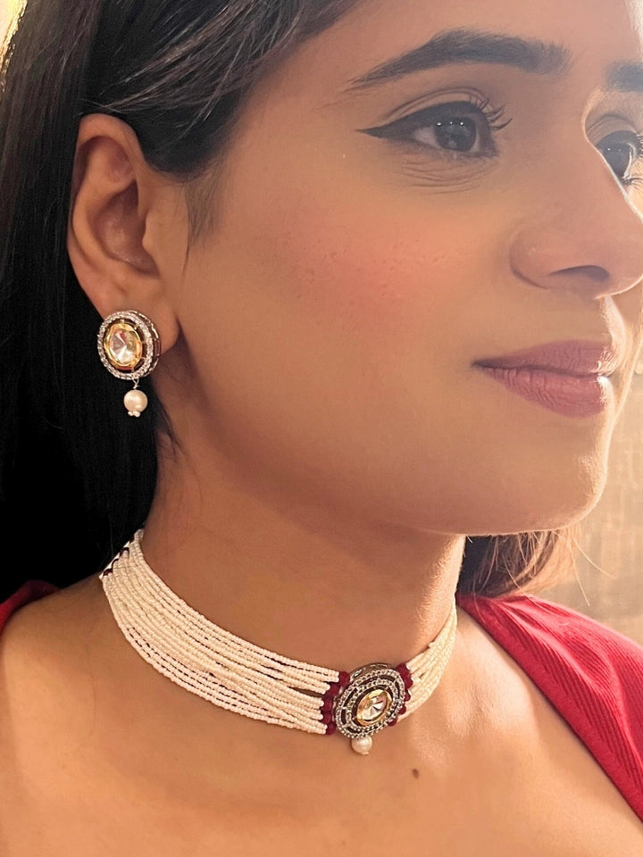 A woman wears the Pearl Red Renu Necklace Set, featuring a multi-strand white pearl choker with Kundan and red accents, a central jeweled pendant, and matching round earrings adorned with dangling pearls. Kundan choker necklace accentuates her look.