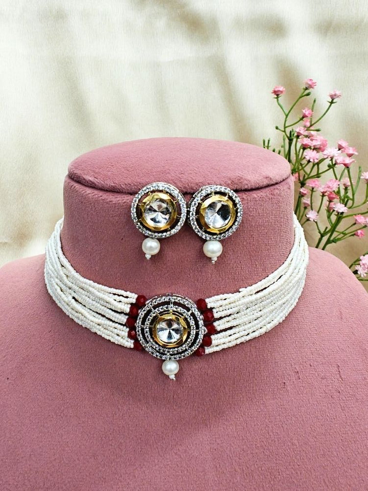 The Pearl Red Renu Necklace Set features a beaded choker with a bold circular pendant and matching stud earrings, embellished with gold, silver, American diamonds, and pearl accents. Displayed on a pink stand with pink flowers in the background.