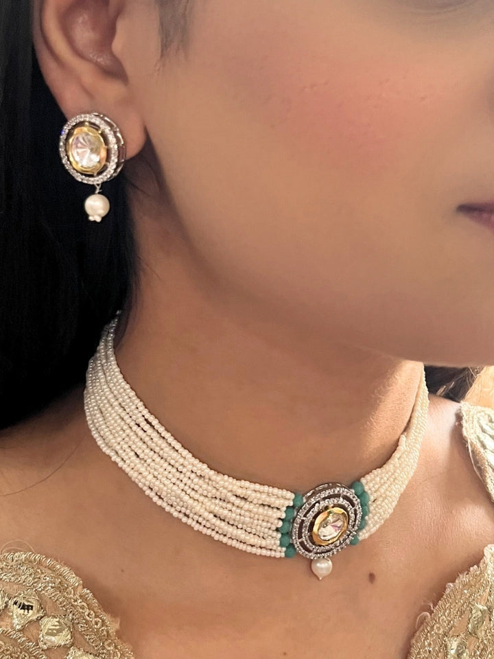 A woman wears the Pearl Teel Renu Necklace Set, featuring a multi-strand white pearl choker with Kundan and Teel accents, a central jeweled pendant, and matching round earrings adorned with dangling pearls. Kundan choker necklace accentuates her look.