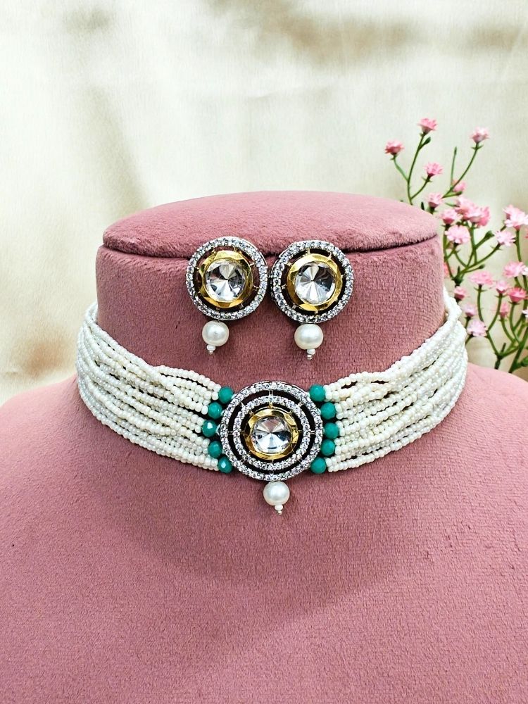 The Pearl Teel Renu Necklace Set features a beaded choker with a bold circular pendant and matching stud earrings, embellished with gold, silver, American diamonds, and pearl accents. Displayed on a pink stand with pink flowers in the background.